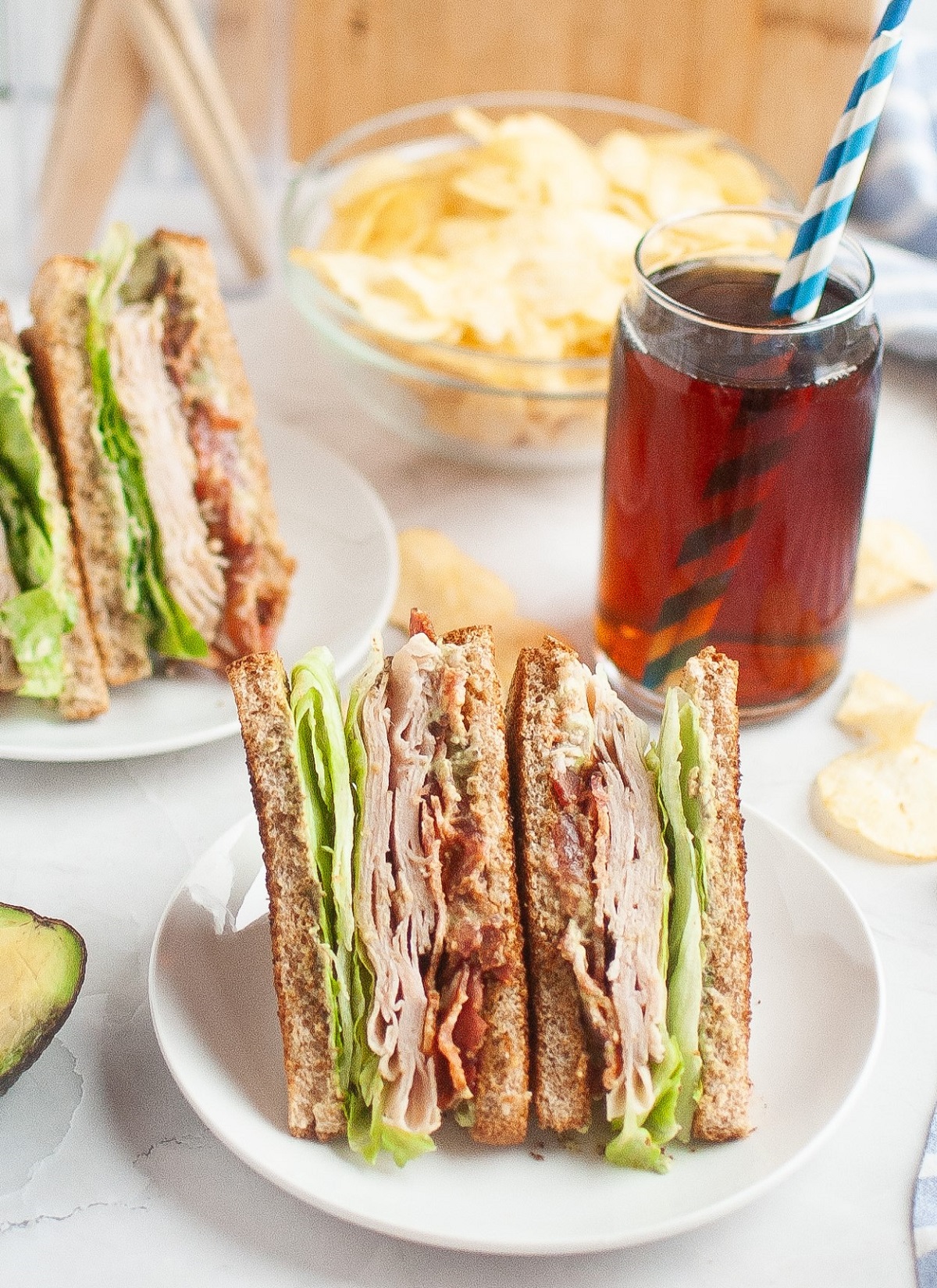 Sliced turkey blt sandwiches on a plate with chips and a glass of tea in the background. 