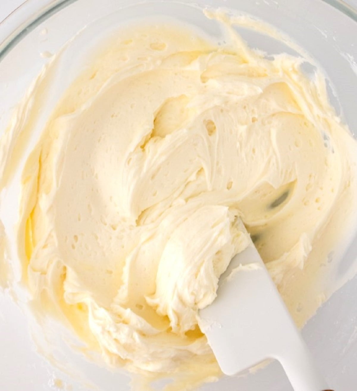 A white spatula stirs fluffy vanilla frosting in a clear bowl.