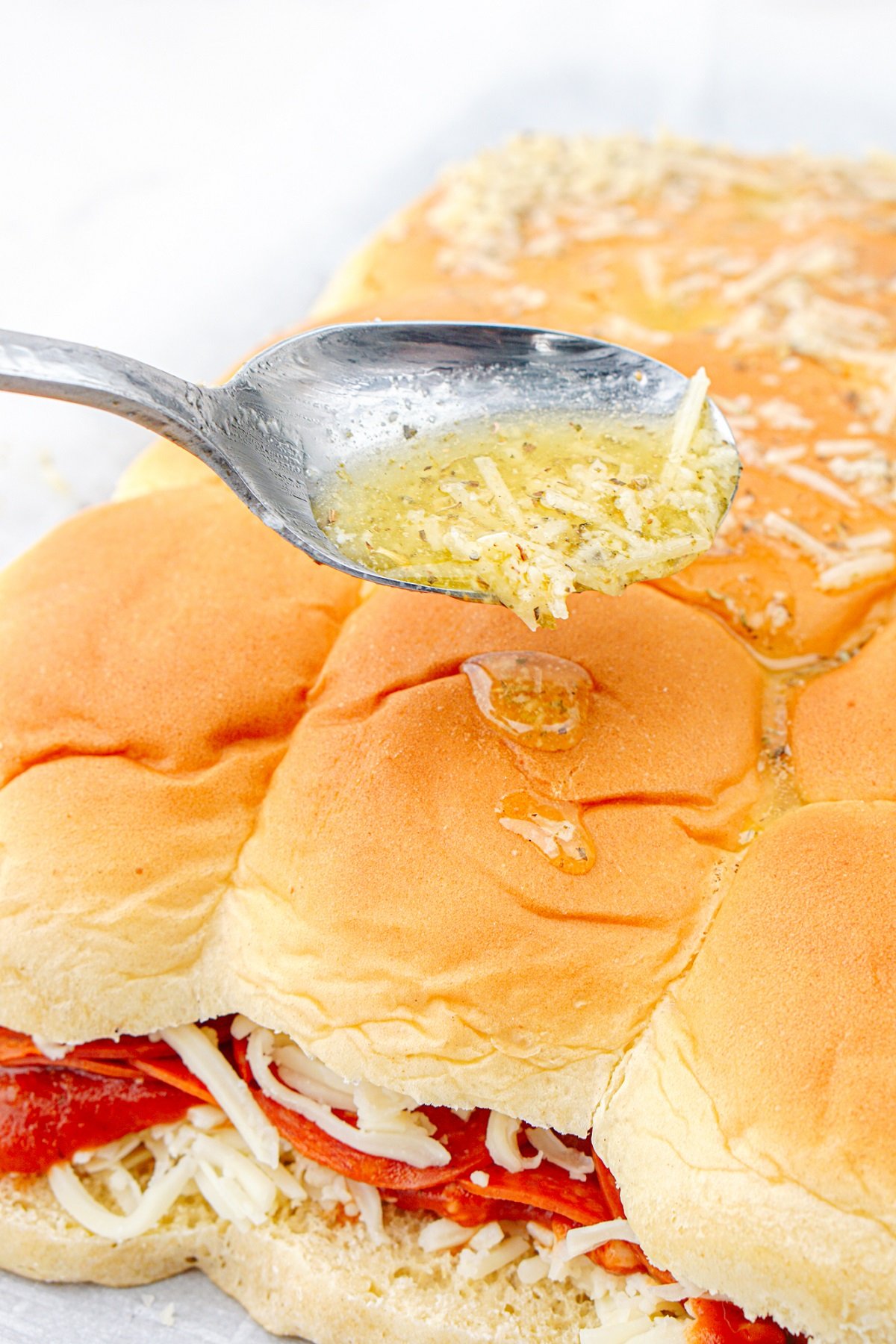 A spoon drizzles garlic butter over the top of Hawaiian rolls. 