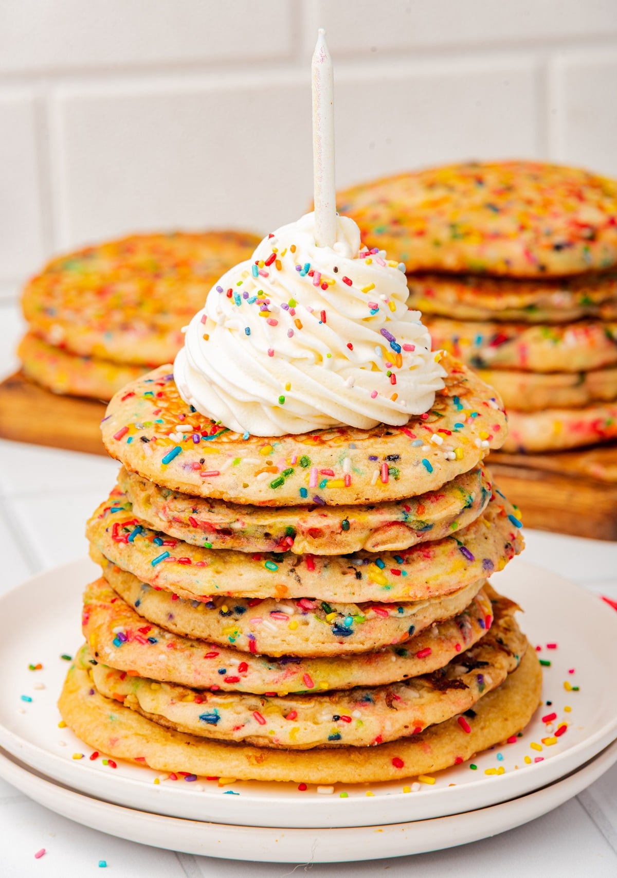 A stack of funfetti pancakes on a plate topped with whipped cream and a birthday candle. 