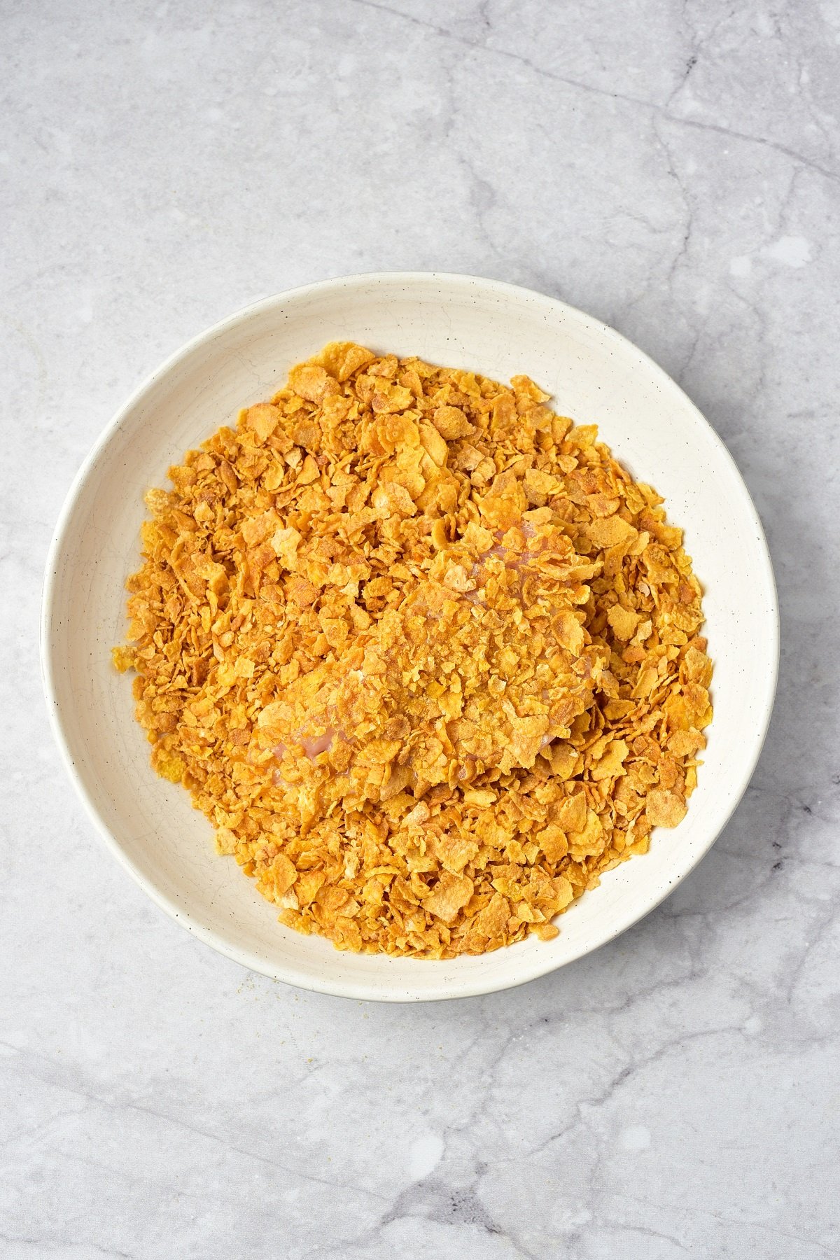 Chicken breast is coated in crushed cornflakes in a small bowl.