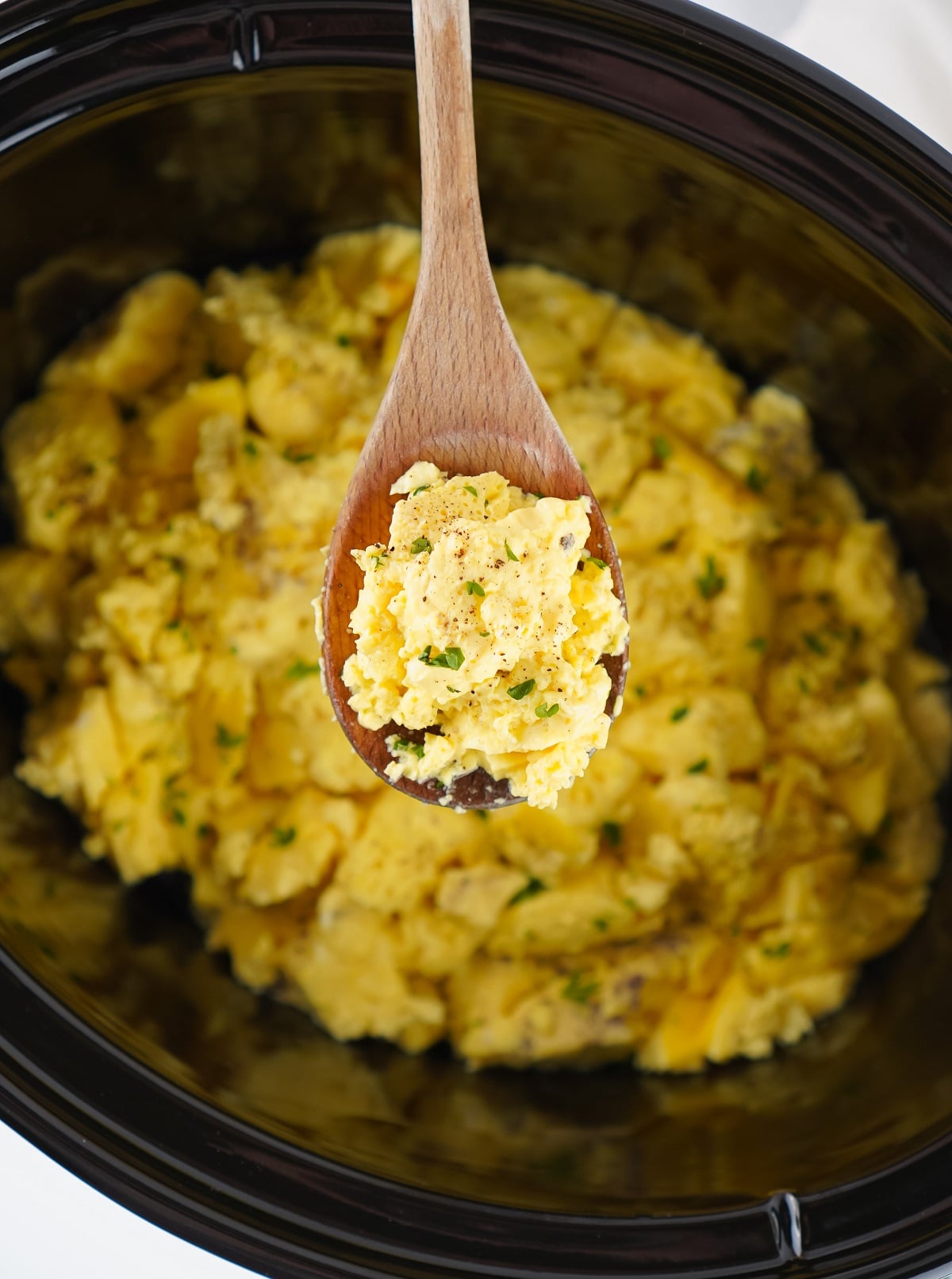 A wooden spoon serves fluffy scrambled eggs from a slow cooker. 