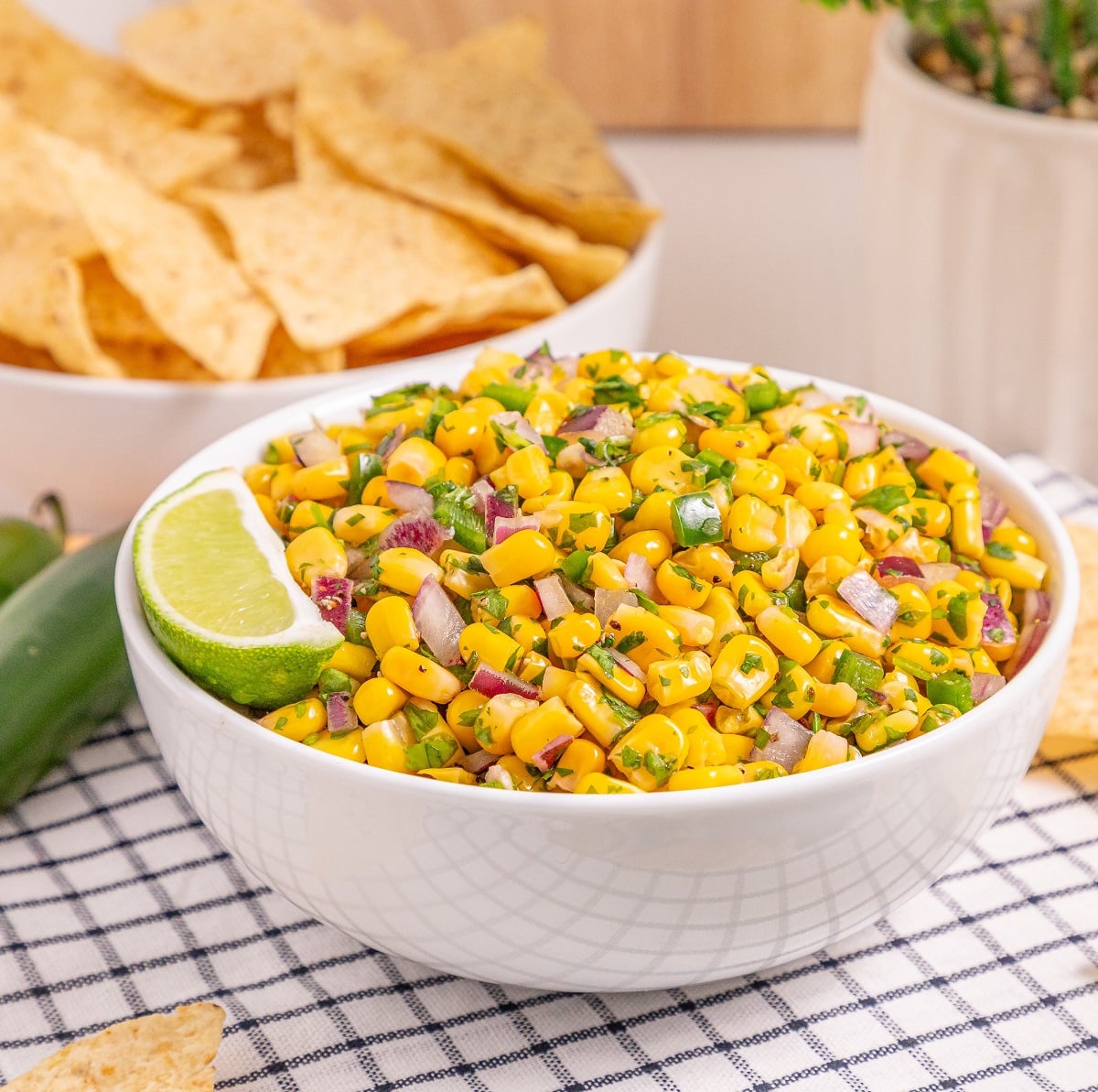 A large white bowl of corn salsa garnished with lime and tortilla chips. 